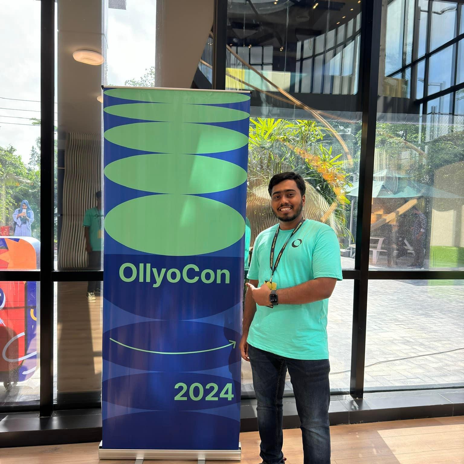 Ofi Khan is standing beside an Ollyocon banner at Ollyocon 2024